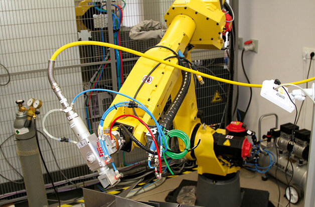 Ogólny widok zrobotyzowanego stanowiska do spawania i cięcia laserowego w Laboratorium Robotyki Wydziału Mechatroniki i Lotnictwa Wojskowej Akademii Technicznej. W skład stanowiska wchodzi sześcioosiowy robot firmy FANUC wyposażony w głowicę do spawania/cięcia laserowego oraz dwuosiowy pozycjoner
